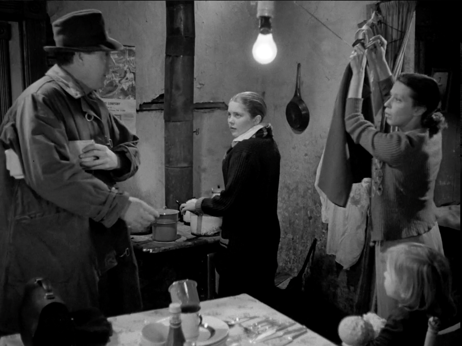 Black and white still. Three white individuals, one man, one child and one woman stand around a table looking at each other. 