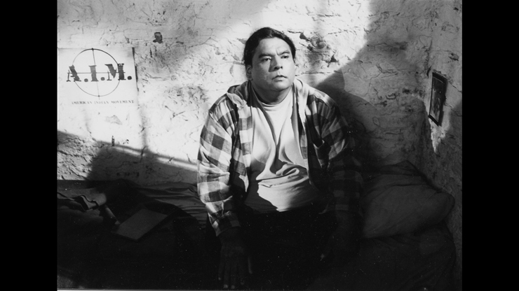 A black and white image of a person sitting on the ground in a plaid shirt, looking beyond the camera. There is a poster in the background that says A.I.M.